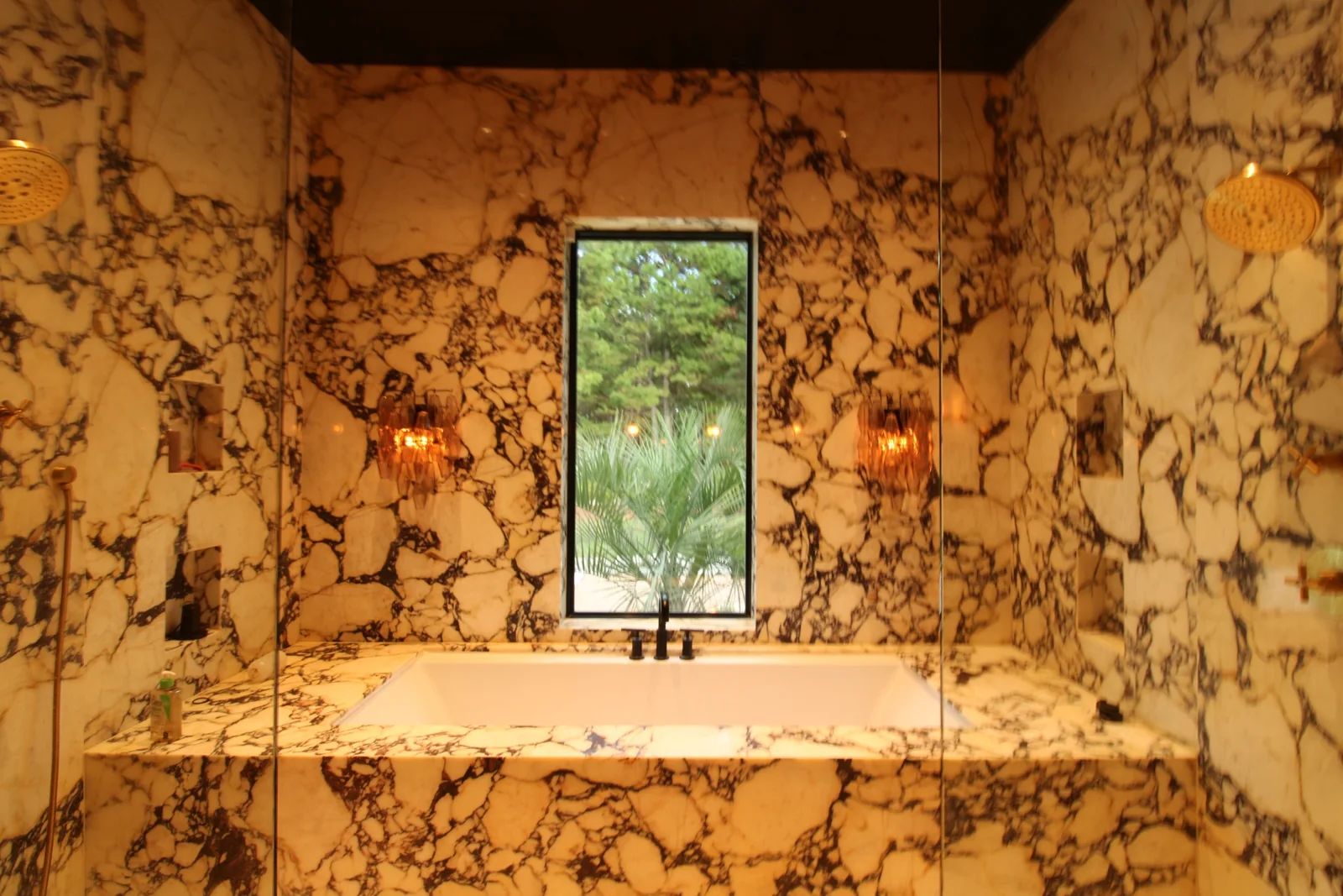 Bathtub set into richly veined stone-patterned surrounds beneath a tall window, with warm wall sconces