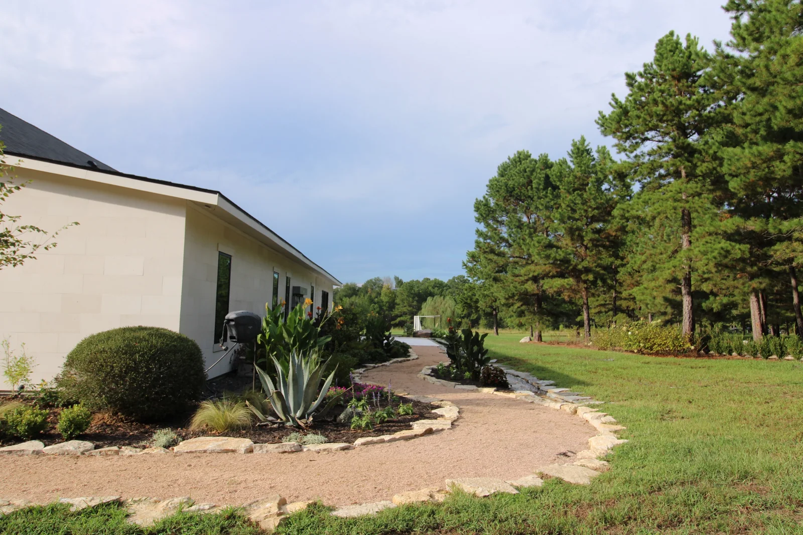 Pale exterior wall, clipped shrubs, and stone-edged gravel beds alongside the ranch home