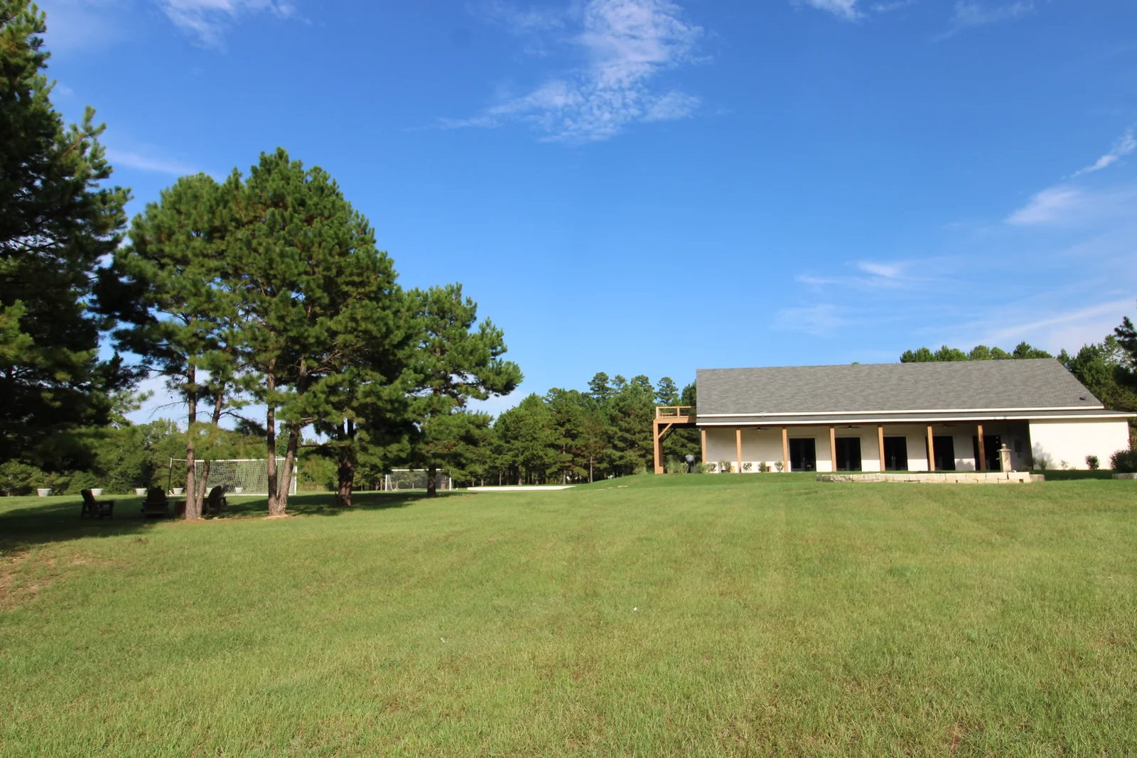 The long porch of the pale ranch home beyond a broad green lawn, with pine trees and blue sky