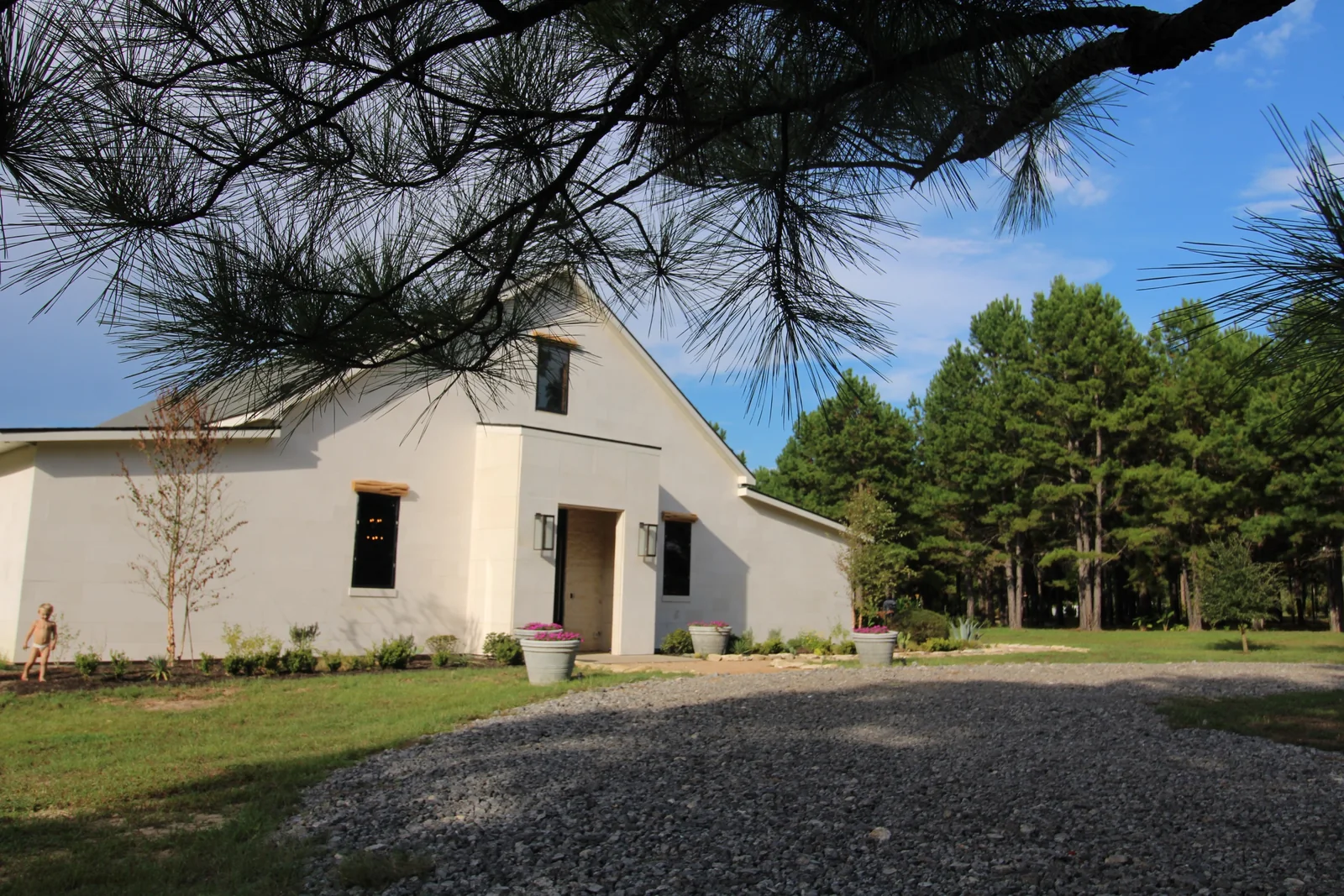 A gabled side of the ranch house under pine branches, beside the gravel drive